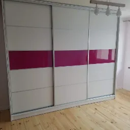Trzydrzwiowa szafa przesuwna z białymi i jasnoszarymi panelami, akcentowana poziomym pasem w kolorze fuksji, na tle drewnianej podłogi i okna