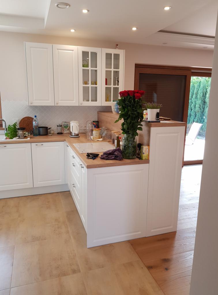 Biała kuchnia z drewnianym blatem, wyspą i bukietem czerwonych róż w wazonie. Widoczne białe szafki wiszące i stojące, płytki na ścianie oraz okno z widokiem na ogród.