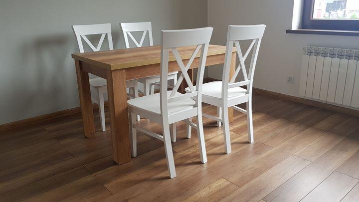 Drewniany stół z jasnym blatem i czterema białymi krzesłami z oparciami w kształcie litery X, ustawiony na podłodze z paneli w ciepłym odcieniu brązu, z grzejnikiem widocznym w tle.