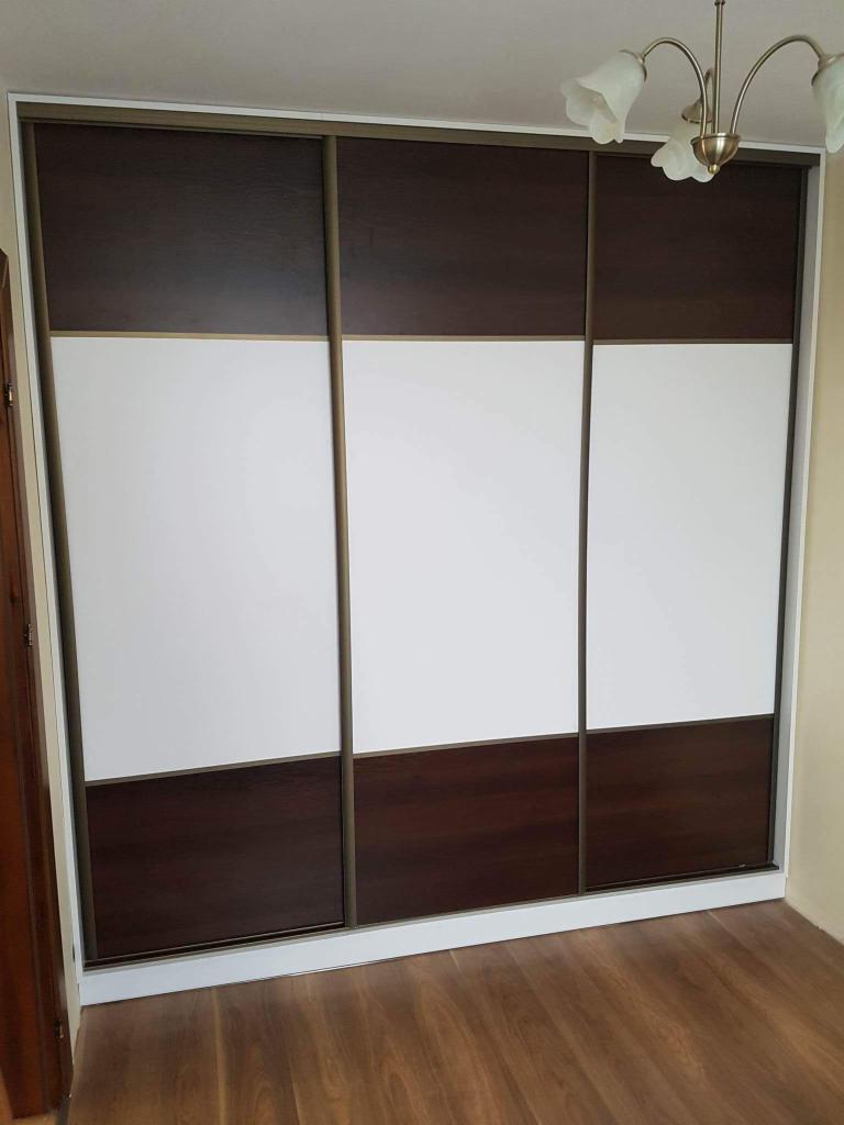 Trzydrzwiowa szafa przesuwna z ciemnobrązowymi i białymi panelami, widoczna lampa sufitowa z kloszami w kształcie kwiatów.