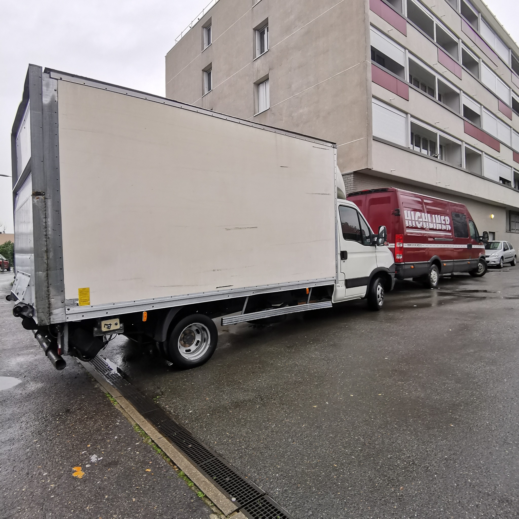 Biała ciężarówka dostawcza zaparkowana na mokrej ulicy miejskiej, w tle budynek mieszkalny i czerwony bus z napisem 'HIGHLINER'.