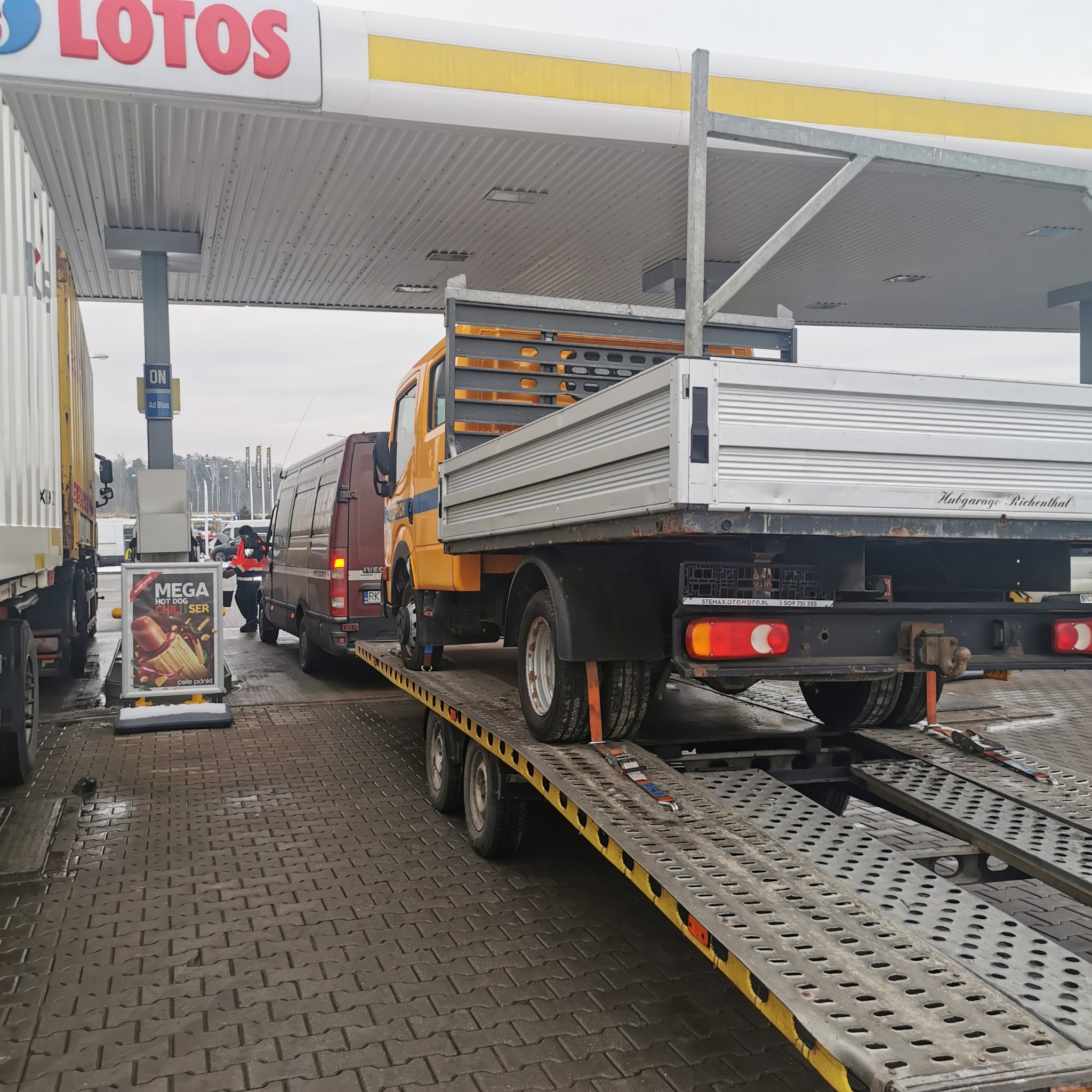 Żółta ciężarówka Iveco z otwartą platformą załadunkową, transportowana na lawecie pod zadaszeniem stacji benzynowej Lotos, widoczny van Iveco i reklama Mega Hot Dog Chilliser.