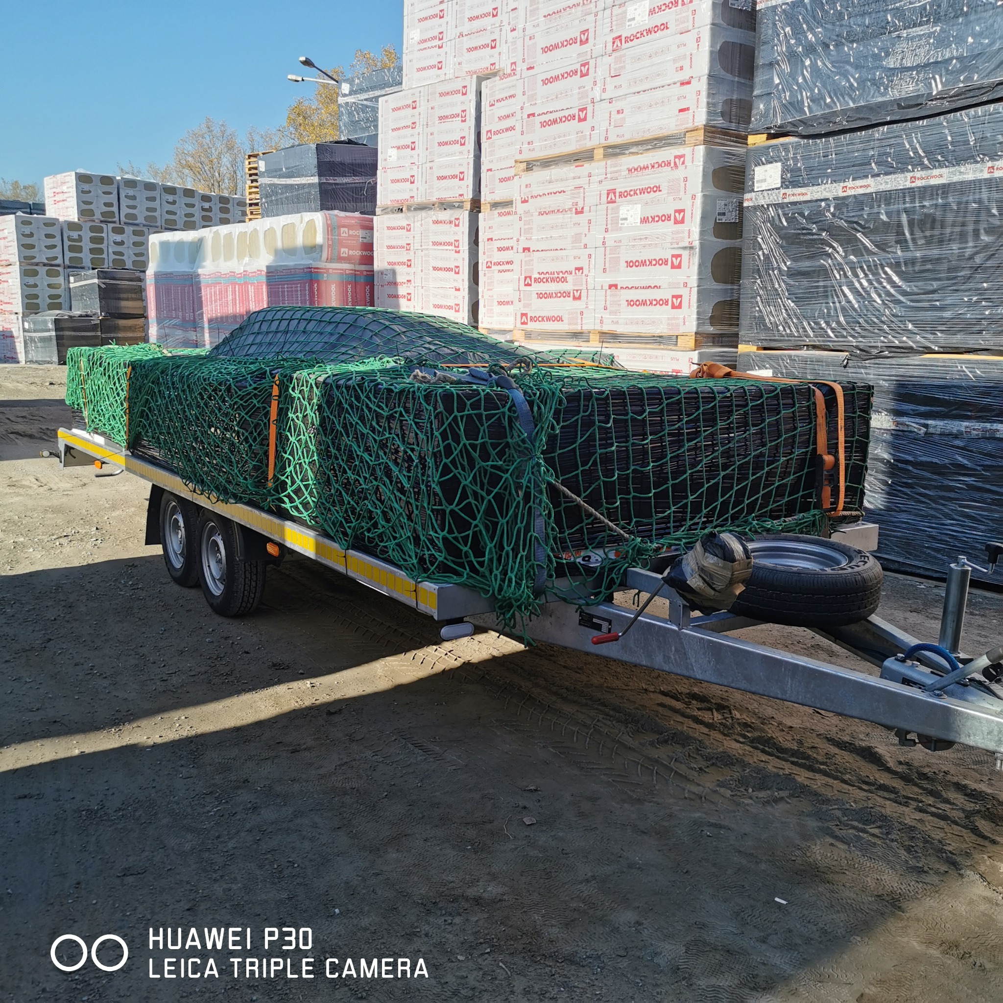Przyczepa samochodowa z zabezpieczonym ładunkiem wełny mineralnej Rockwool, zapakowanej w folię, na tle magazynu z paletami materiałów budowlanych, w słoneczny dzień.