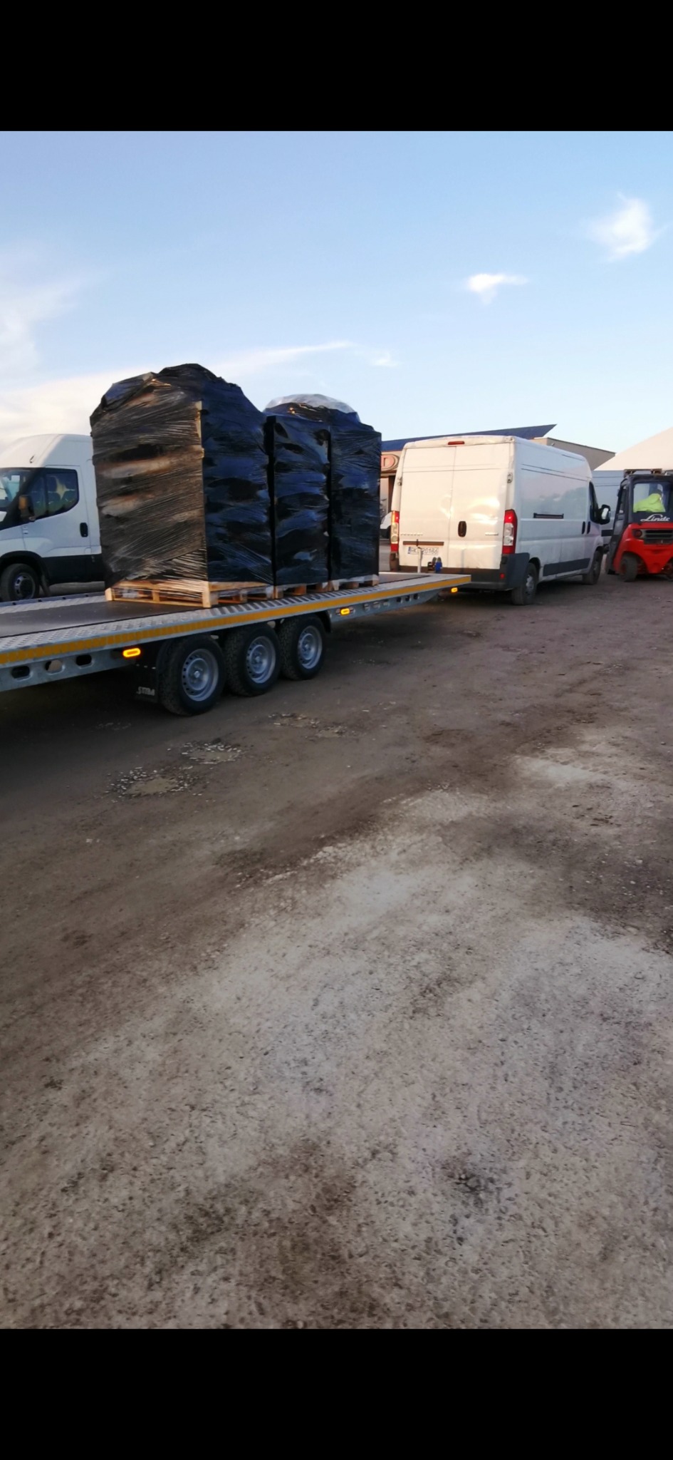 Przyczepa z trzema dużymi, owiniętymi w czarną folię ładunkami na paletach, ciągnięta przez biały bus, z widocznym w tle wózkiem widłowym i innymi pojazdami.