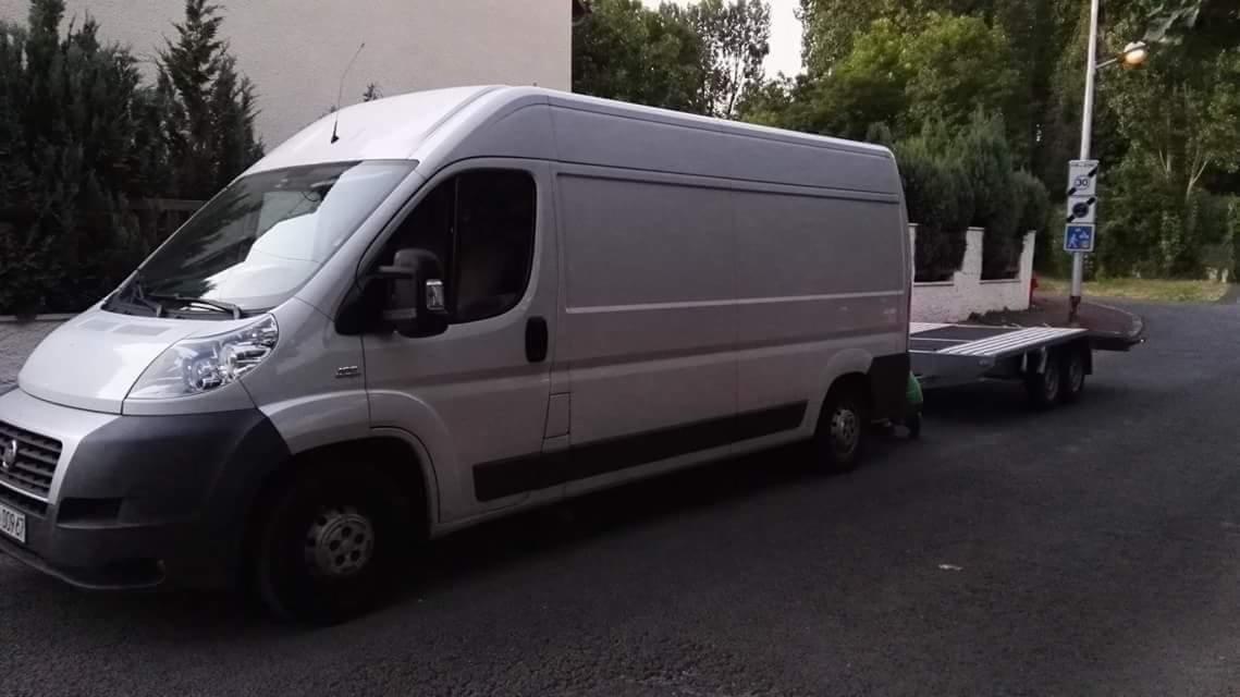 Laweta na haku za busem Fiat Ducato na tle ulicy i zieleni. Bus w kolorze białym, laweta dwuosiowa. Tablica rejestracyjna busa widoczna.