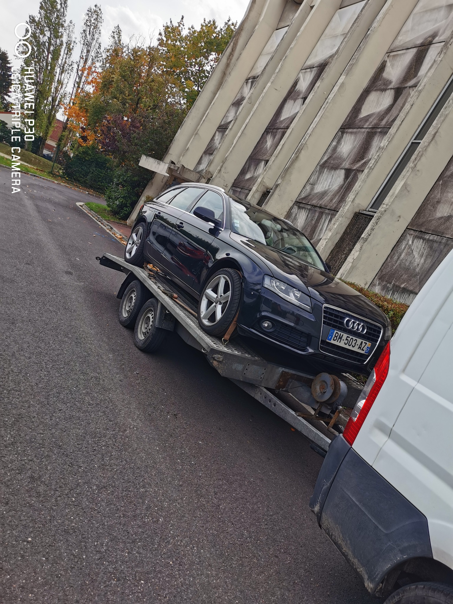 Czarny samochód Audi A4 z francuską tablicą rejestracyjną BM-503-AZ transportowany na niskopodwoziowej przyczepie za białym busem na tle modernistycznego budynku.