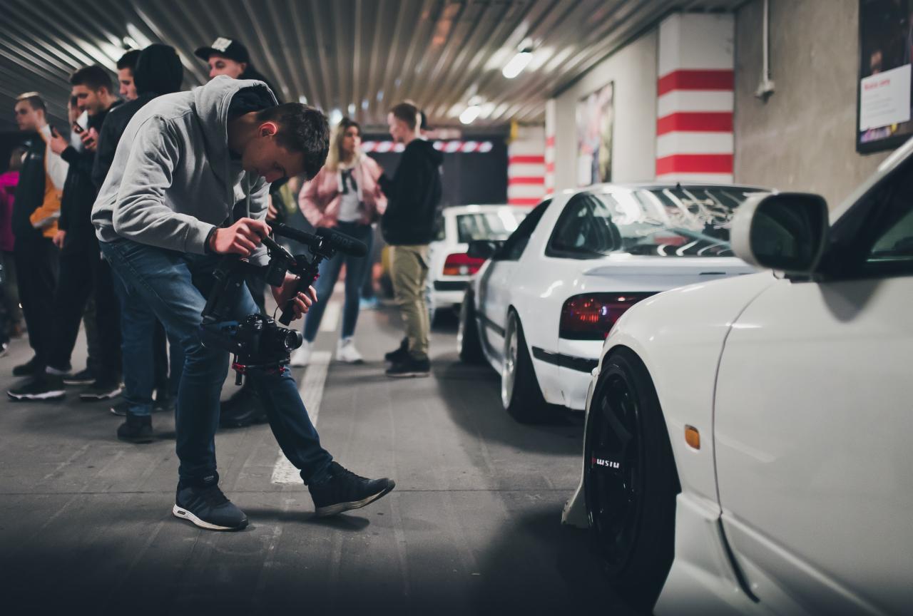 Młody mężczyzna z profesjonalną kamerą filmuje białe samochody sportowe w garażu podziemnym, w tle grupa osób.