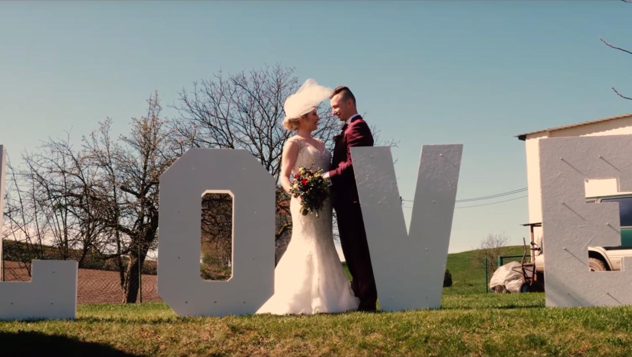 Para młoda obejmująca się za literami LOVE na tle wiejskiego krajobrazu w słoneczny dzień. Panna młoda w koronkowej sukni, pan młody w bordowym garniturze.