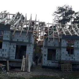 Dwa budynki w stanie surowym otwartym z szarego bloczka, widoczne drewniane elementy konstrukcji dachu, w tle drzewa i sylwetka człowieka.