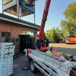 Czerwony podnośnik koszowy przy sklepie Netto, robotnik w koszu na wysokości montuje szyld. Na przyczepie drabiny i akcesoria. Stosy worków na palecie.