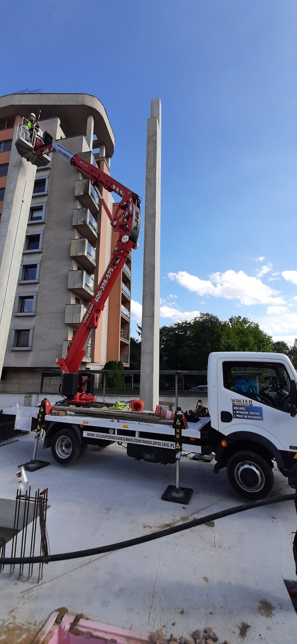 Czerwony podnośnik koszowy na białym aucie, na wysokości budynku. Pracownik w koszu podnośnika. Błękitne niebo w tle. Widoczne logo firmy na aucie.