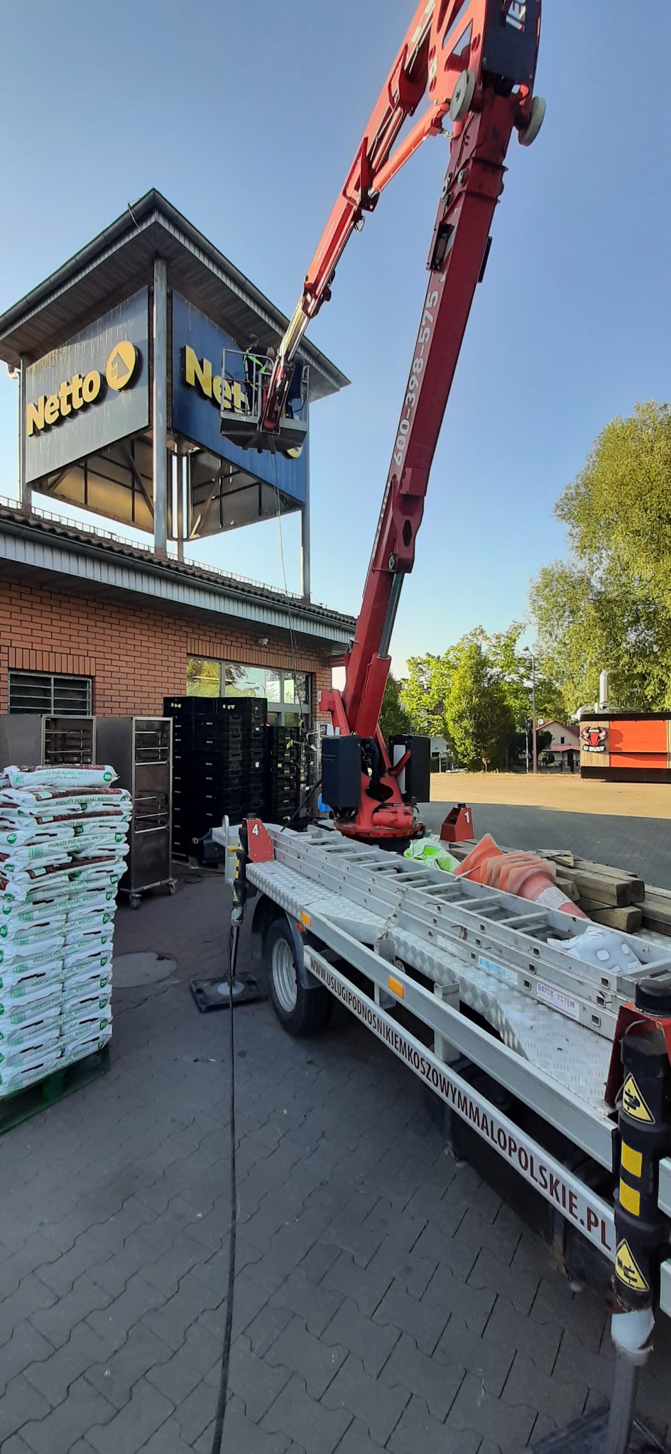 Czerwony podnośnik koszowy przy sklepie Netto, robotnik w koszu na wysokości montuje szyld. Na przyczepie drabiny i akcesoria. Stosy worków na palecie.