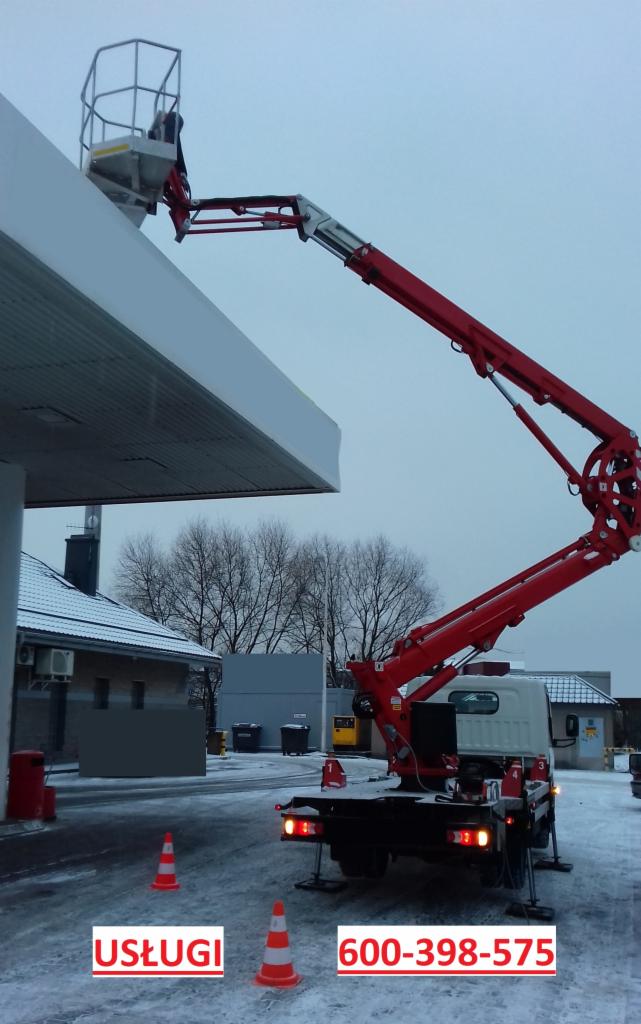 Czerwony podnośnik koszowy przy białym zadaszeniu stacji benzynowej, widoczne pomarańczowe pachołki drogowe i numer telefonu kontaktowego.