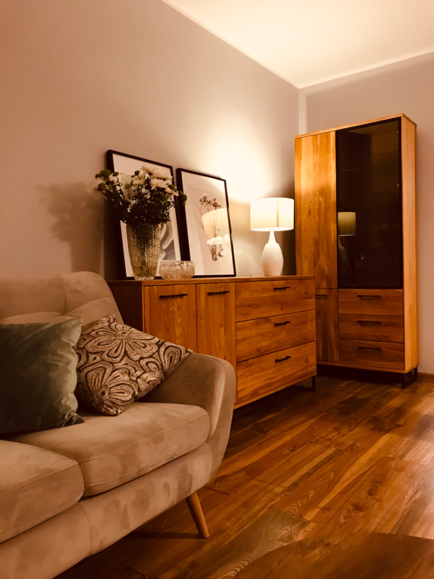 Jasny salon z drewnianą komodą, witryną i beżową sofą z dekoracyjnymi poduszkami, oświetlony lampą stołową i naturalnym światłem.