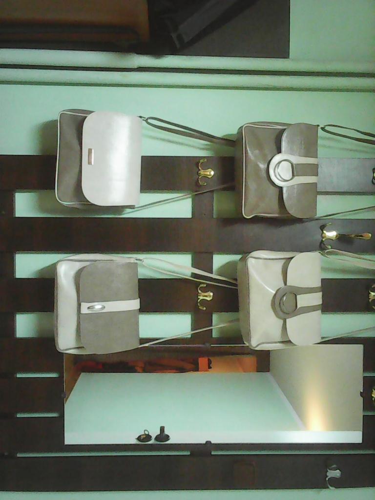 Cztery torebki damskie w odcieniach beżu i szarości, wiszące na ozdobnym, ciemnobrązowym wieszaku z metalowymi uchwytami w kształcie stylizowanych kwiatów, na tle jasnej, zielonkawej ściany.