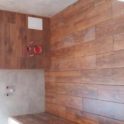 Remonty - Ściana łazienki wyłożona brązowymi płytkami imitującymi drewno, z widocznymi przyłączami hydraulicznymi i białymi elementami armatury w tle, kontrastującymi z szarymi płytkami na pozostałej części...