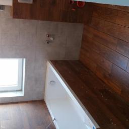 Remonty - Wnętrze łazienki po remoncie, widoczne płytki imitujące drewno na podłodze i ścianie, biała wanna z folią ochronną, szare płytki na ścianie z oknem.