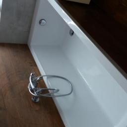 Remonty - Nowoczesna, biała wanna prostokątna z chromowaną baterią, ułożona na podłodze z płytek imitujących drewno, z widocznym fragmentem okna z rośliną doniczkową.