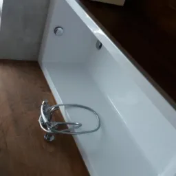 Nowoczesna, biała wanna prostokątna z chromowaną baterią, ułożona na podłodze z płytek imitujących drewno, z widocznym fragmentem okna z rośliną doniczkową.