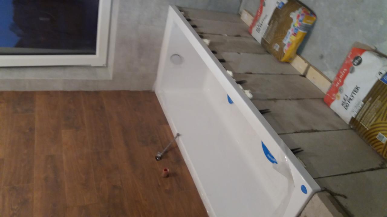 Biały brodzik w trakcie montażu w łazience, widoczne worki z klejem do płytek, drewnopodobne panele podłogowe i fragment okna.