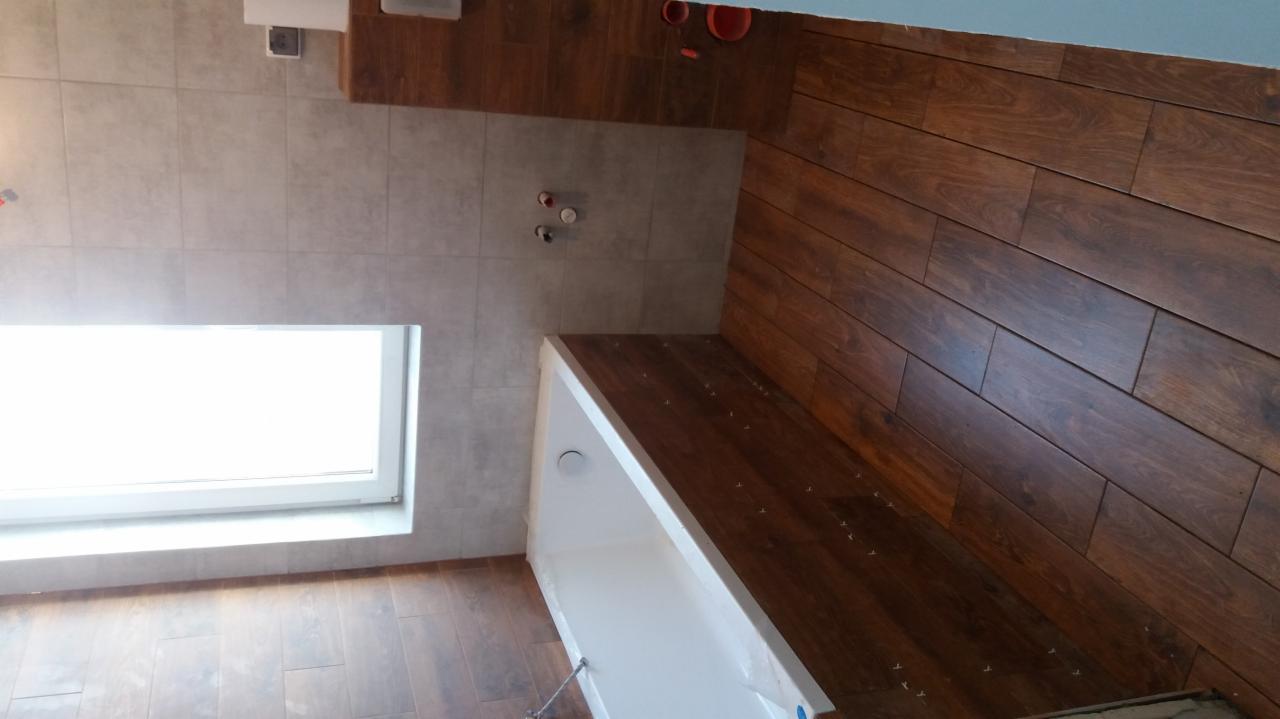 Wnętrze łazienki po remoncie, widoczne płytki imitujące drewno na podłodze i ścianie, biała wanna z folią ochronną, szare płytki na ścianie z oknem.