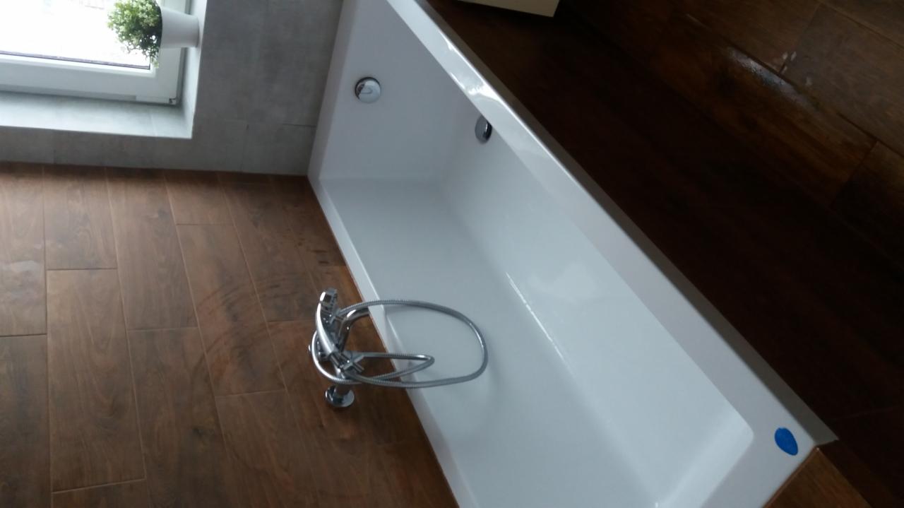 Nowoczesna, biała wanna prostokątna z chromowaną baterią, ułożona na podłodze z płytek imitujących drewno, z widocznym fragmentem okna z rośliną doniczkową.