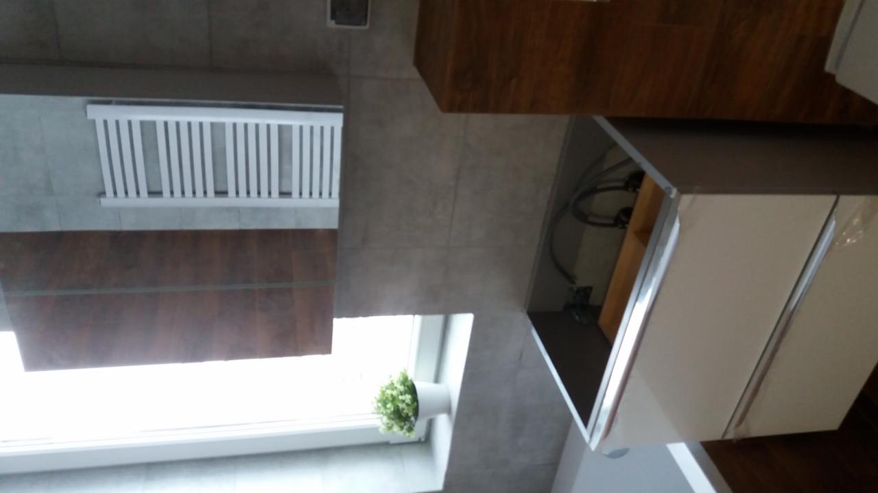 Ukończona łazienka z szarymi płytkami, białym grzejnikiem drabinkowym, drewnianą szafką pod umywalką i białą szafką z umywalką, oraz doniczką z kwiatami na parapecie okna.