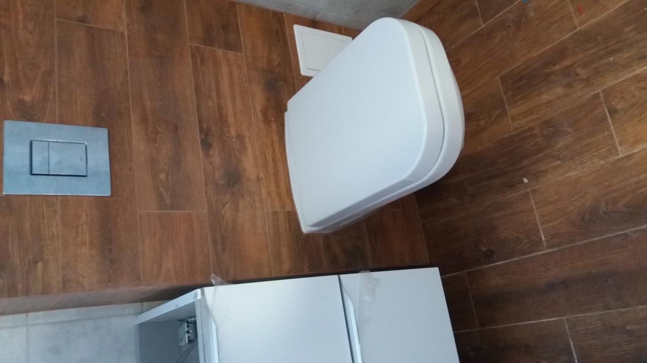 Wisząca toaleta w nowoczesnej łazience z brązowymi płytkami imitującymi drewno, srebrny przycisk spłuczki i biała szafka pod umywalką z folią ochronną.