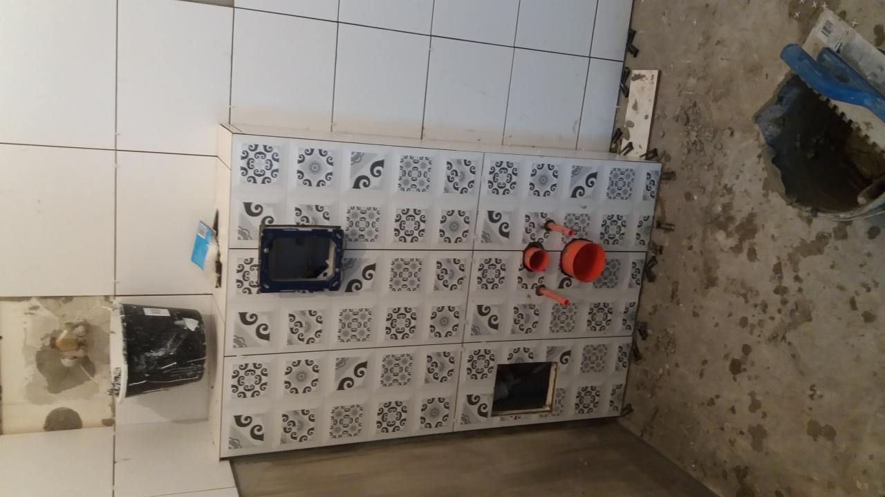 Wnętrze łazienki w trakcie prac: Częściowo ułożone białe płytki z dekoracyjnym wzorem na obudowie stelaża WC, widoczne elementy instalacji hydraulicznej i otwory rewizyjne, surowy beton na podłodze.