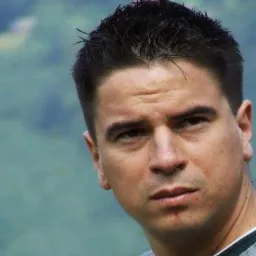 Portret mężczyzny z krótkimi, ciemnymi włosami i poważnym wyrazem twarzy na rozmytym, zielonym tle.