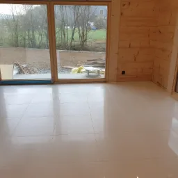 Jasne, błyszczące płytki podłogowe w nowym pomieszczeniu z drewnianymi ścianami i dużymi oknami z widokiem na ogród w tle.