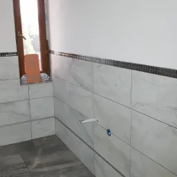 Świeżo wykończona łazienka z szarymi płytkami imitującymi marmur na podłodze i ścianach, dekoracyjna mozaika na górze ścian, widoczny fragment okna z brązową ramą i pomarańczowa packa do fugowania...