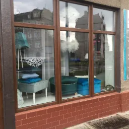 Wystawa sklepu z tkaninami obiciowymi i dekoracyjnymi, prezentująca tapicerowane pufy w odcieniach niebieskiego i zieleni, widoczne przez okno z brązowymi ramami. Na zewnątrz budynku widoczna...