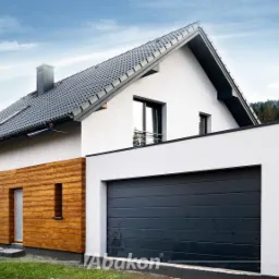 Nowoczesny dom jednorodzinny z białymi ścianami, drewnianą elewacją i grafitową bramą garażową, widok z zewnątrz w słoneczny dzień.