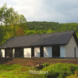 Parterowy dom z płaskim dachem krytym ciemną dachówką, z dużymi oknami i białymi ścianami, na tle zielonego wzgórza i drzew, w słoneczny dzień.