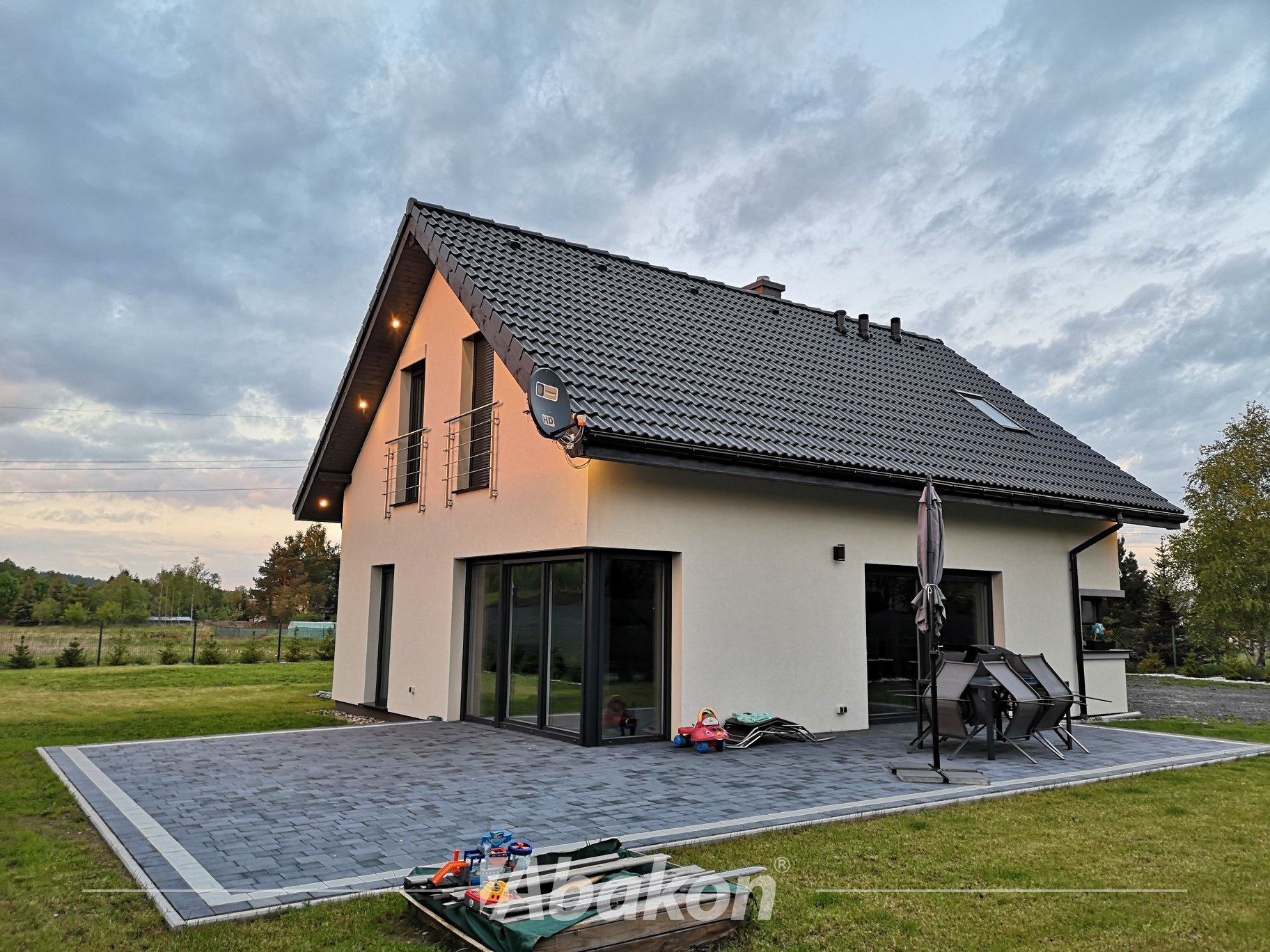 Nowoczesny dom jednorodzinny z jasną elewacją, grafitowym dachem i dużym tarasem z kostki brukowej; widoczne zabawki dziecięce i meble ogrodowe.