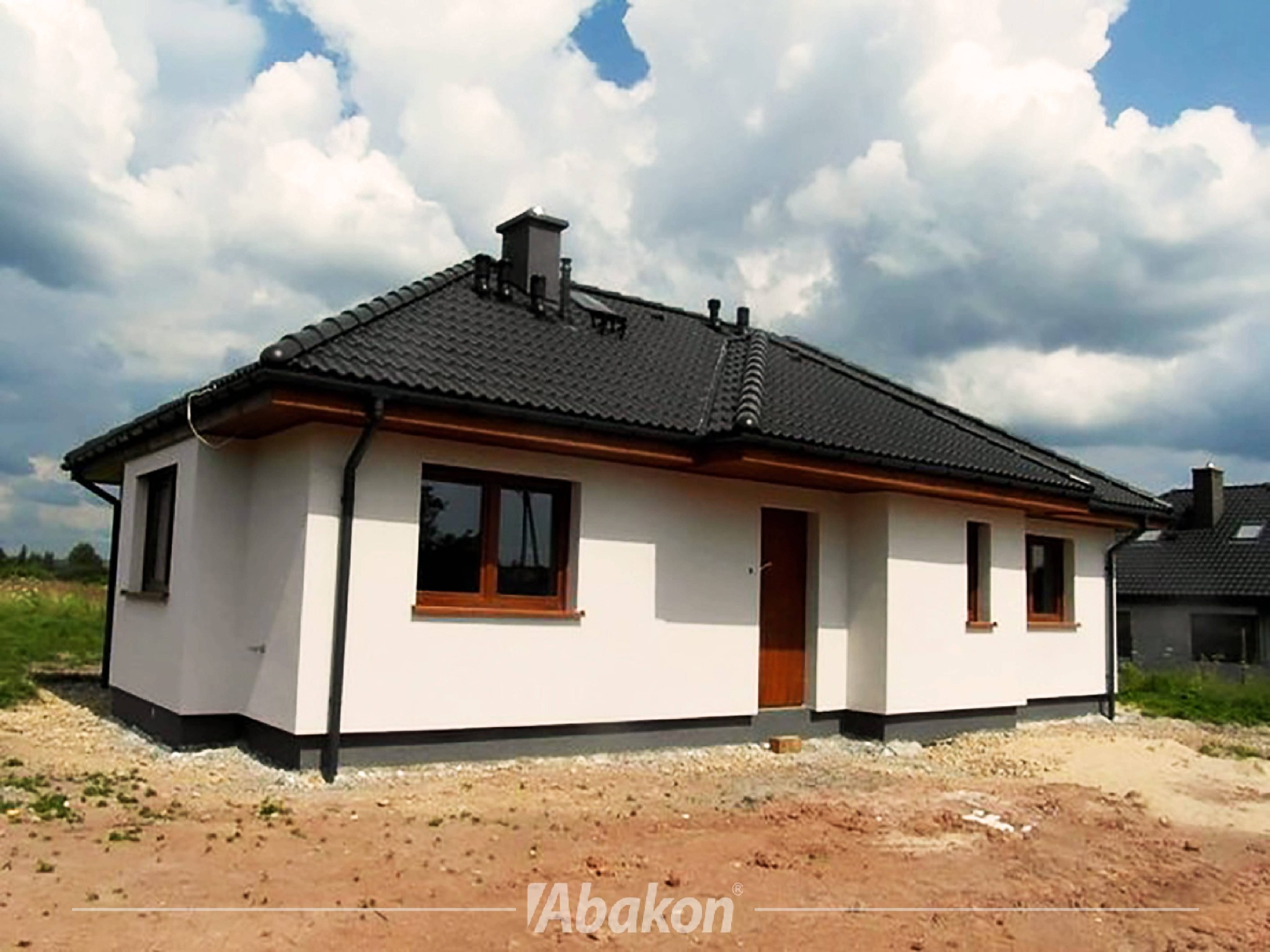 Parterowy dom z białymi ścianami i ciemnobrązowymi oknami, z ciemną dachówką i kominem, na działce z ziemią, pod błękitnym niebem z chmurami.