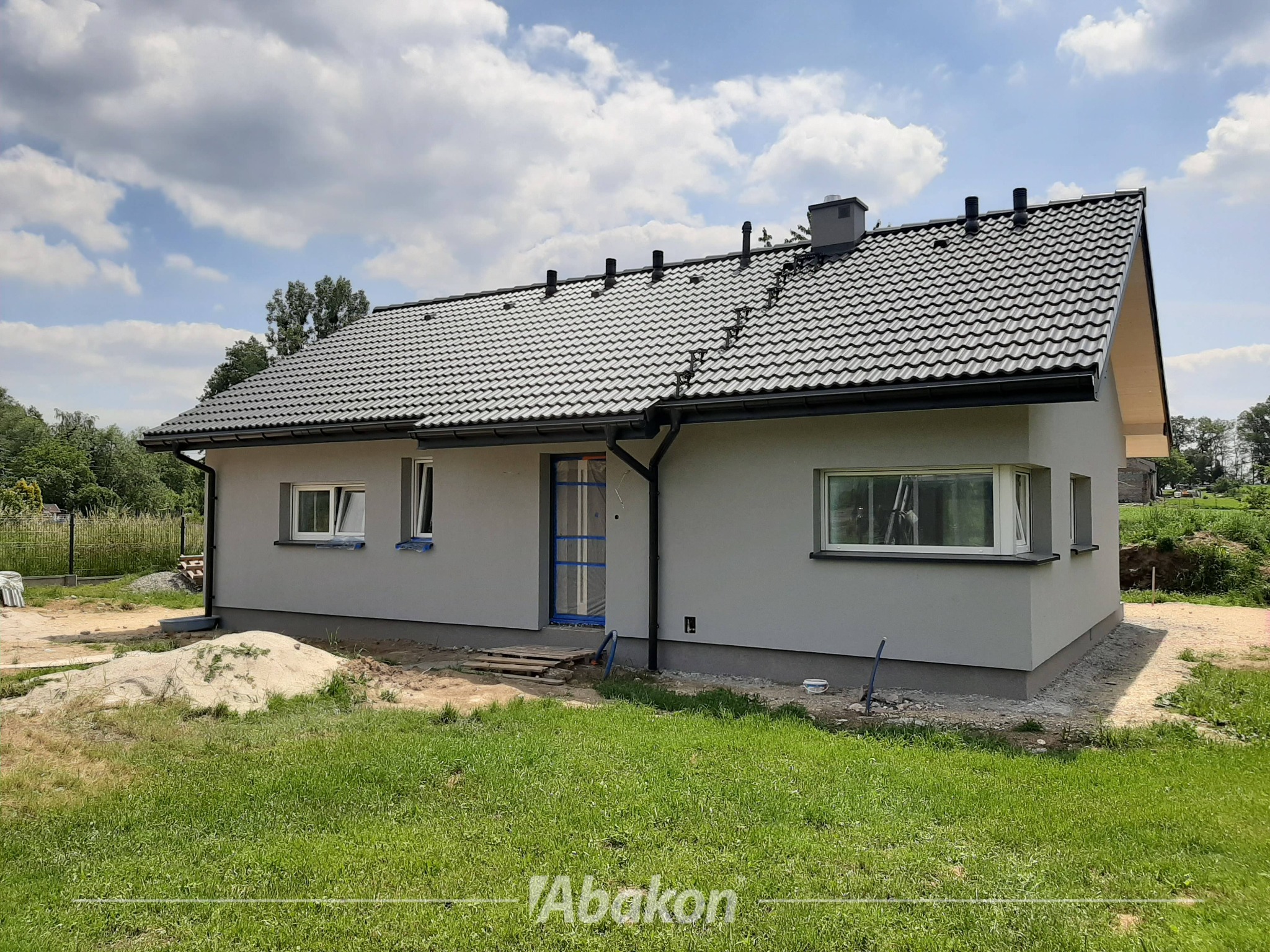 Niski, parterowy dom z szarą elewacją i ciemną dachówką, z widocznymi elementami wykończeniowymi wokół okien i drzwi, na tle błękitnego nieba z chmurami.