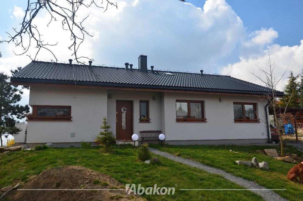 Parterowy dom z białymi ścianami, ciemną dachówką i drewnianymi ramami okiennymi, otoczony trawnikiem i młodymi drzewkami.