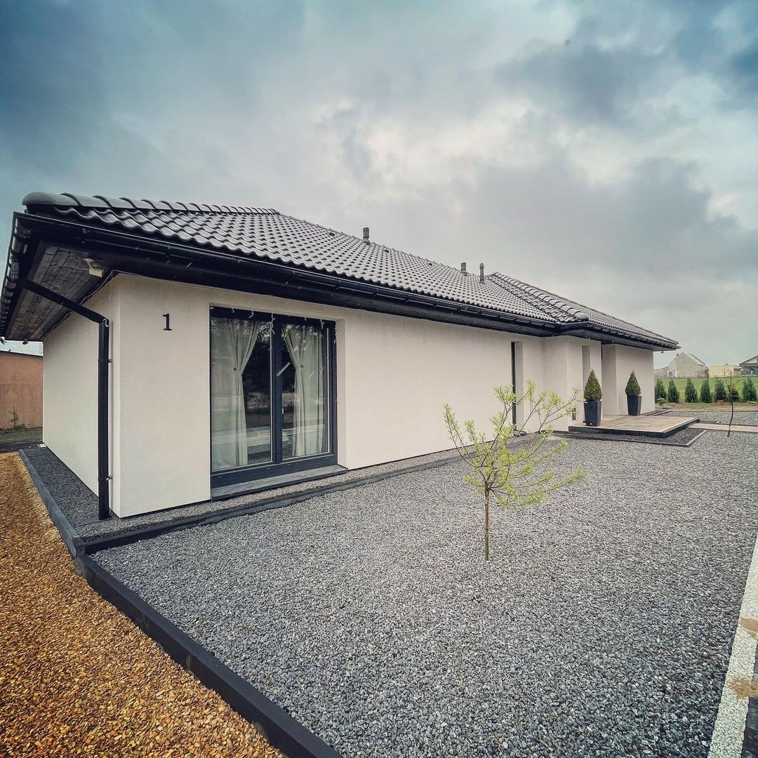 Nowoczesny, parterowy dom z białymi ścianami i ciemną dachówką, numer '1' nad oknem, otoczony szarym żwirem i młodym drzewkiem, widok na pochmurne niebo.