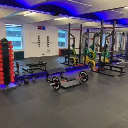 Nowoczesna siłownia z profesjonalnym sprzętem do ćwiczeń siłowych: stojaki na sztangi z obciążeniami, hantle, kettlebells, ławeczki, podłoga wyłożona matą, oświetlenie ledowe.