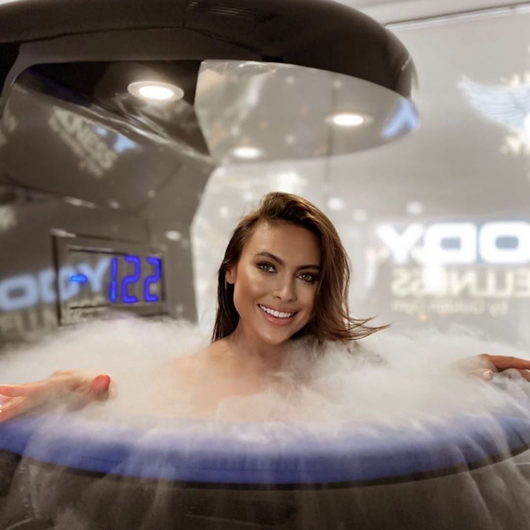 Uśmiechnięta kobieta w kriosaunie otoczona parą, widoczny wyświetlacz z temperaturą, logo studia fitness w tle.