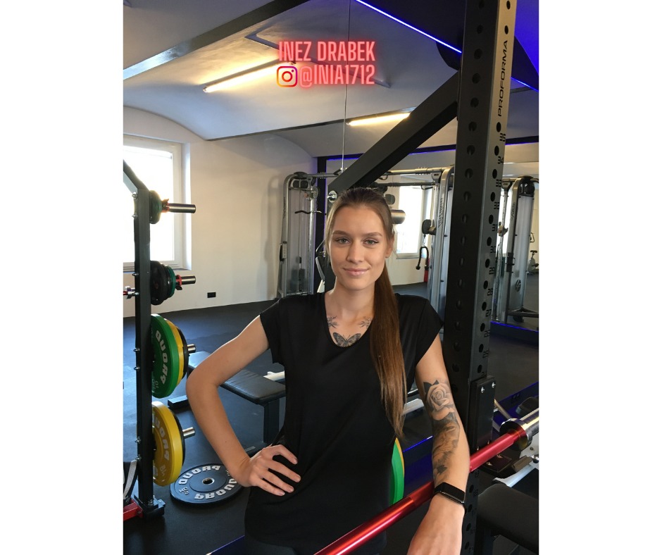 Kobieta z tatuażami, w czarnej koszulce, opiera się o stojak do ćwiczeń z obciążeniami w siłowni. W tle widoczne maszyny do ćwiczeń i okno.