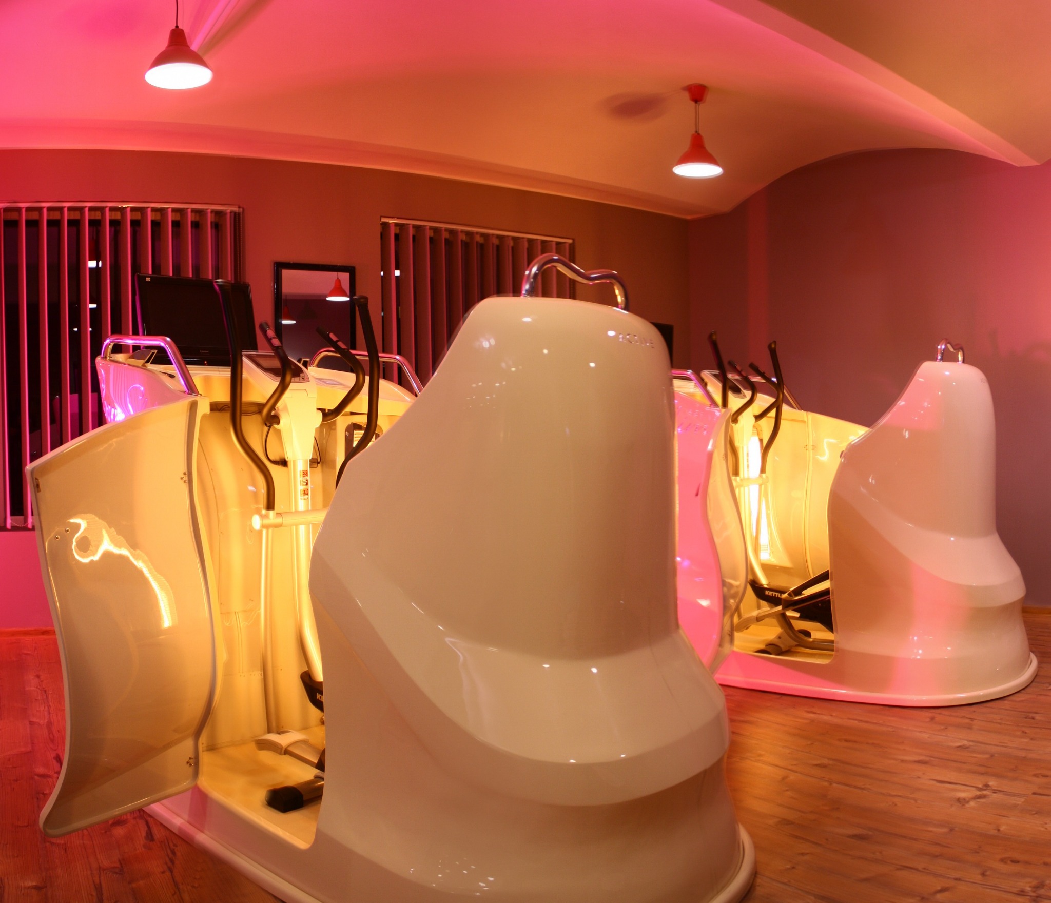 Dwa urządzenia Powerslim Active z podczerwienią w jasnym pomieszczeniu z drewnianą podłogą, widoczne otwarte kapsuły z rowerami eliptycznymi w środku, oświetlone czerwonymi lampami.