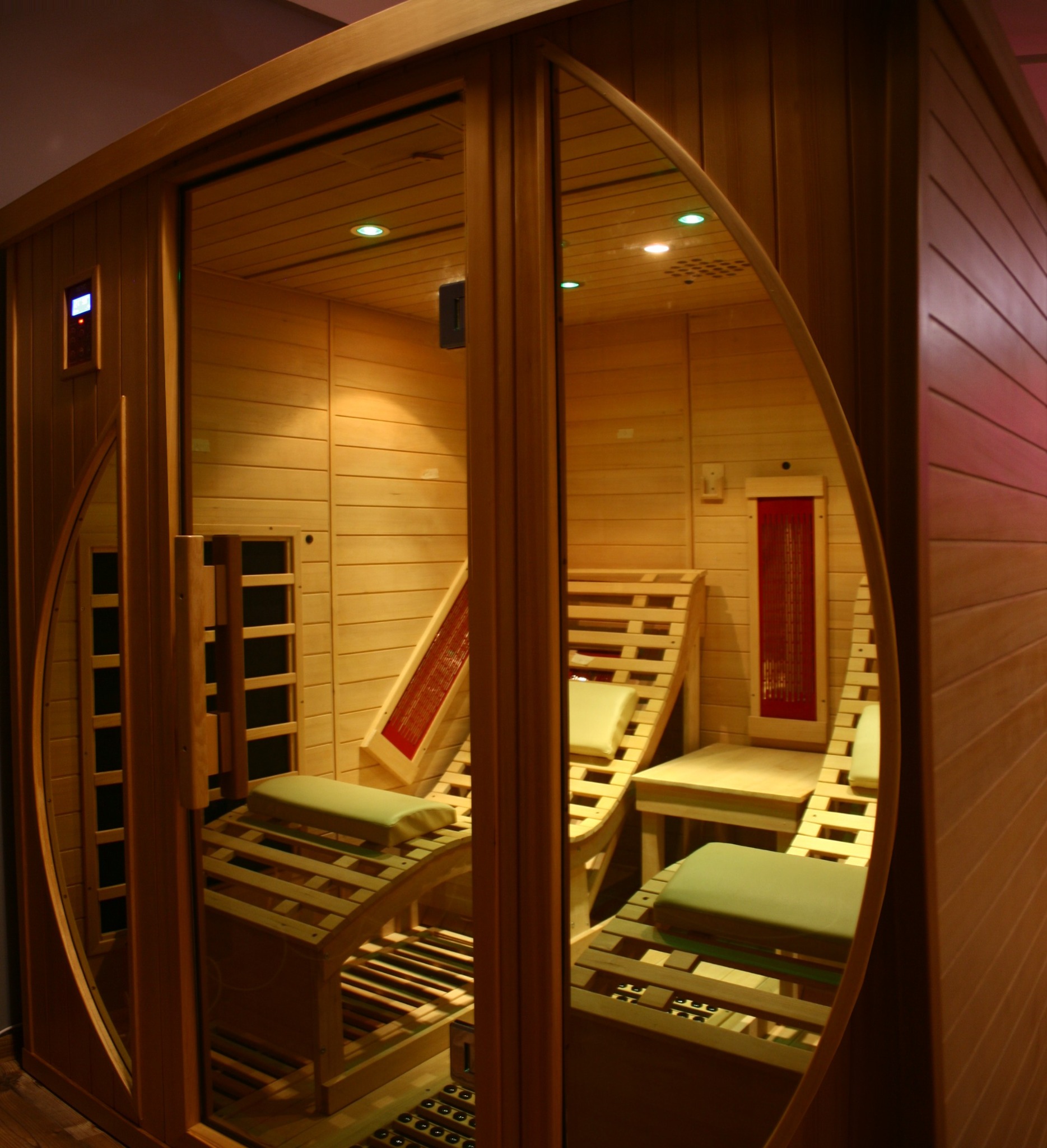 Wnętrze drewnianej sauny infrared z dwoma leżankami z poduszkami, widoczne panele grzewcze na ścianach i sterownik temperatury przy wejściu.