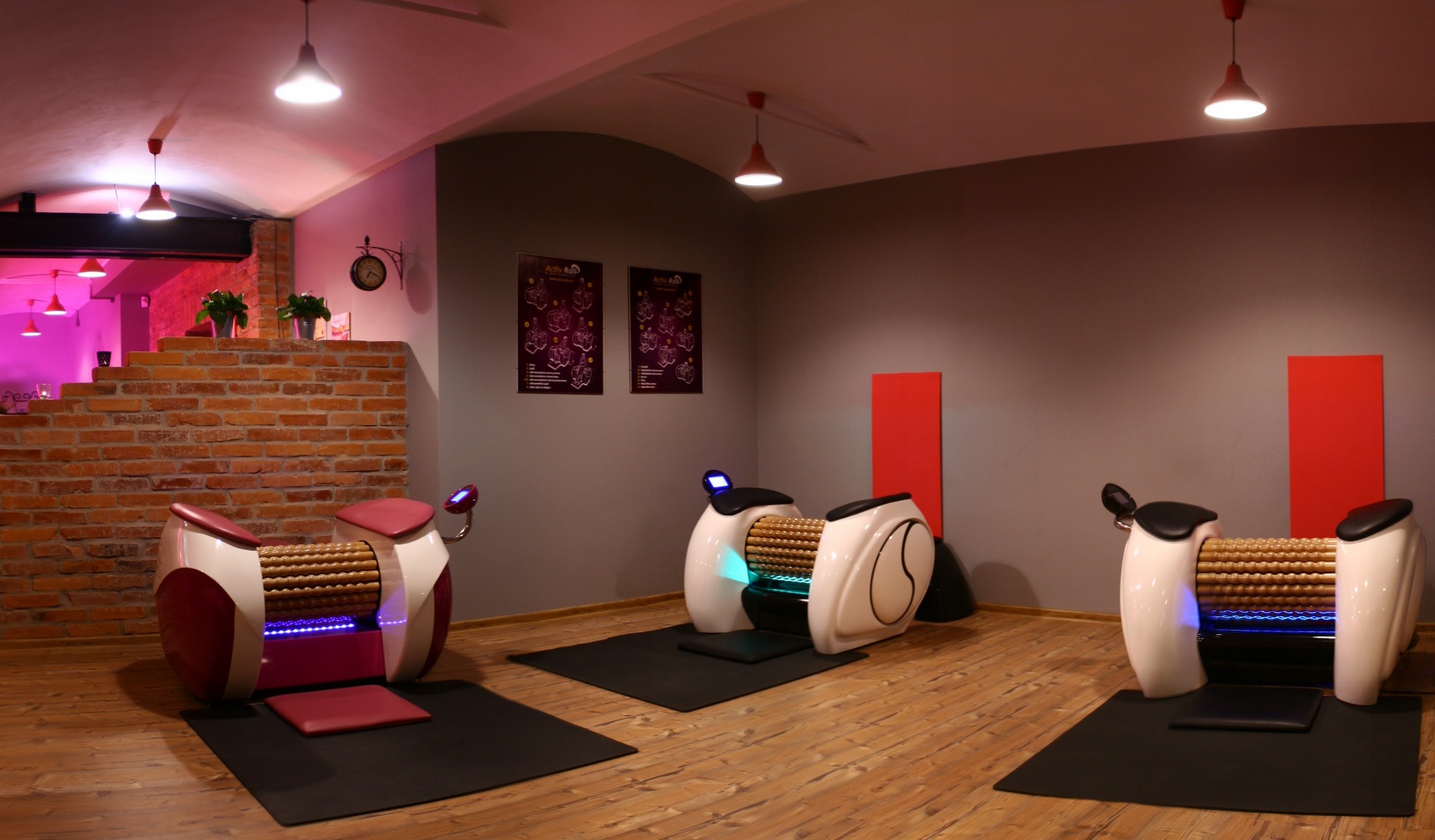 Wnętrze salonu z trzema rollmasażerami z podświetleniem LED na drewnianej podłodze, ściany w odcieniach szarości i cegły, oświetlenie sufitowe.
