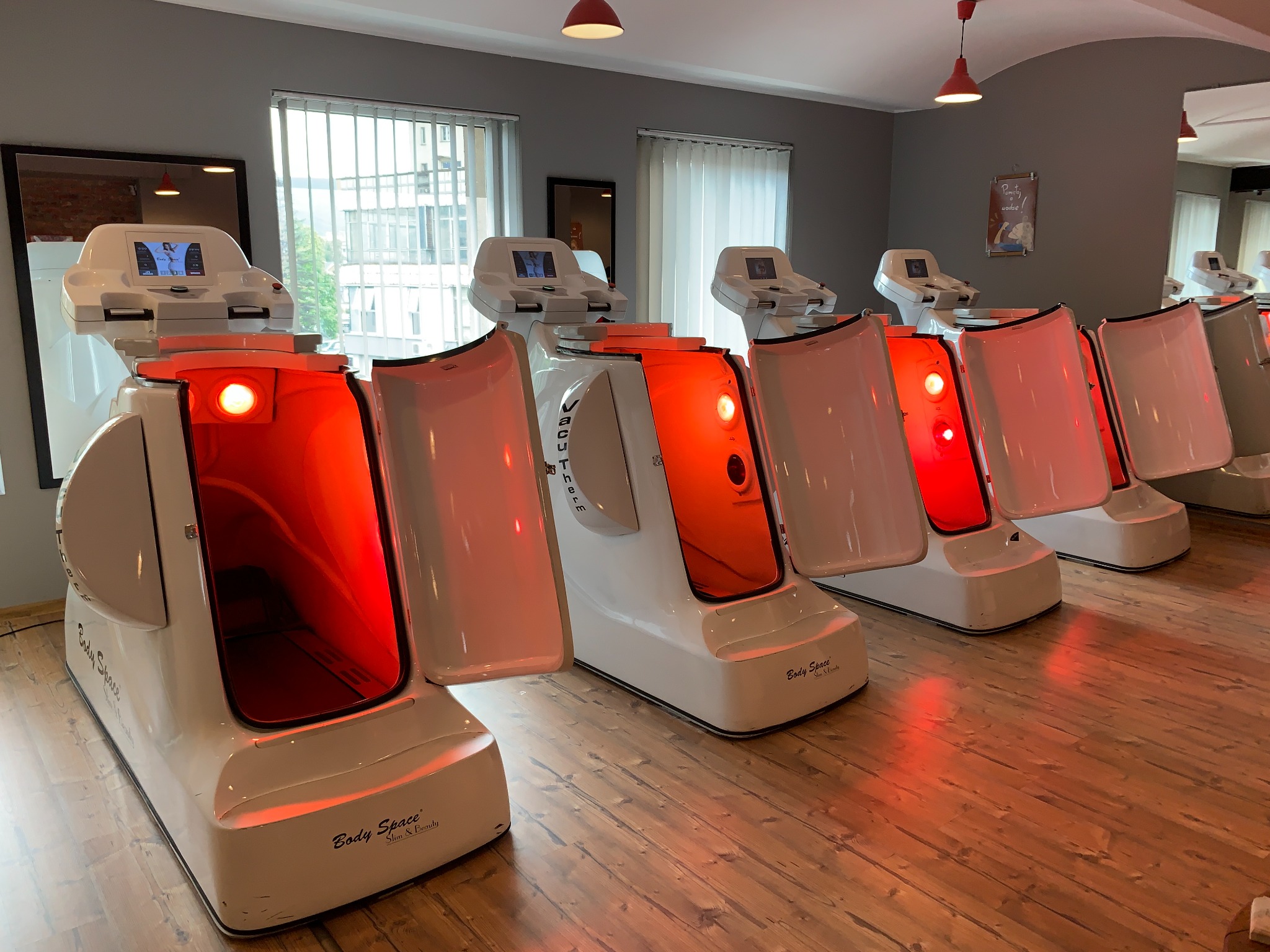 Rząd białych bieżni Vacu Therm Infrared z otwartymi kapsułami i czerwonym światłem wewnątrz, umieszczonych w jasnym pomieszczeniu z drewnianą podłogą.