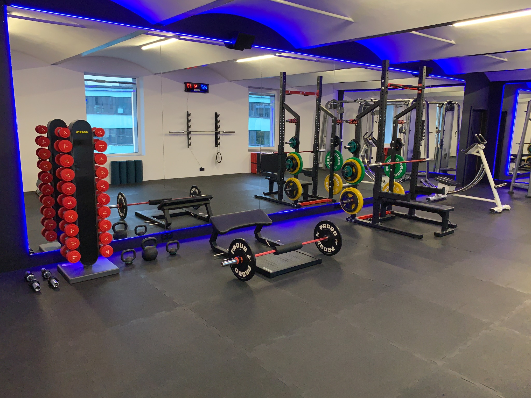 Nowoczesna siłownia z profesjonalnym sprzętem do ćwiczeń siłowych: stojaki na sztangi z obciążeniami, hantle, kettlebells, ławeczki, podłoga wyłożona matą, oświetlenie ledowe.