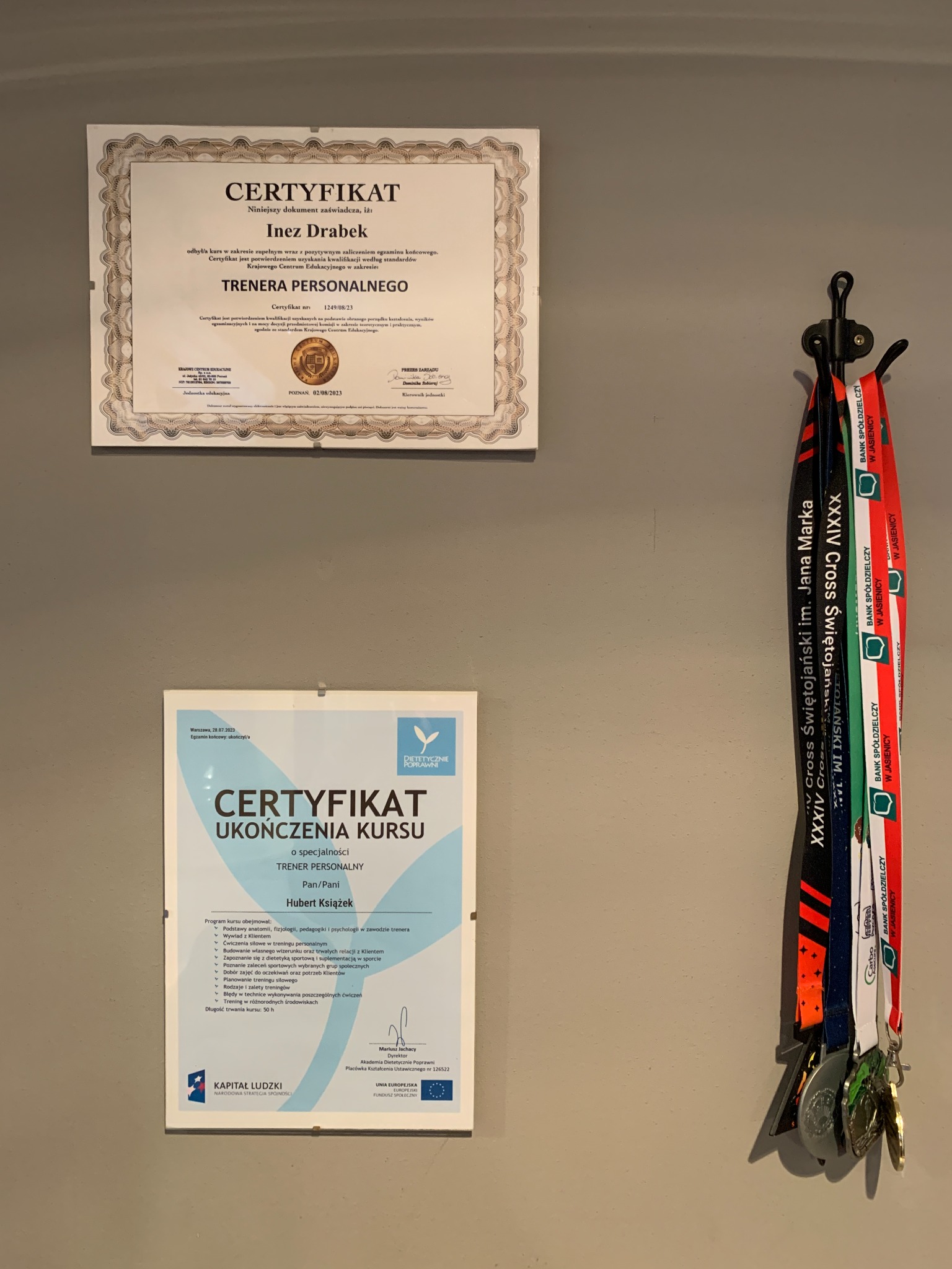 Dwa certyfikaty i medale wiszące na ścianie. Górny certyfikat potwierdza kwalifikacje trenera personalnego Inez Drabek, a dolny certyfikat ukończenie kursu trenera personalnego przez Huberta...
