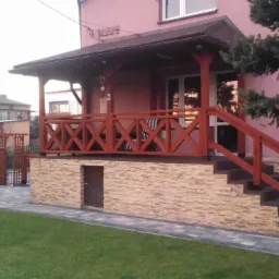 Zadaszone wejście do domu z drewnianą balustradą w kolorze ceglastym, kamienną elewacją i schodami z kostki, otoczone trawnikiem i kostką brukową.